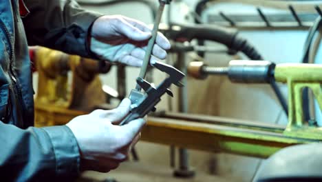 un mecánico en forma de trabajo y guantes mide la altura, el ancho y la longitud de la articulación de bola para un automóvil en el fondo de la sala de servicio.