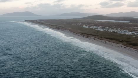 Drone-shot-of-a-couple-walking-on-Berneray-beach,-with-the-machair-in-the-background