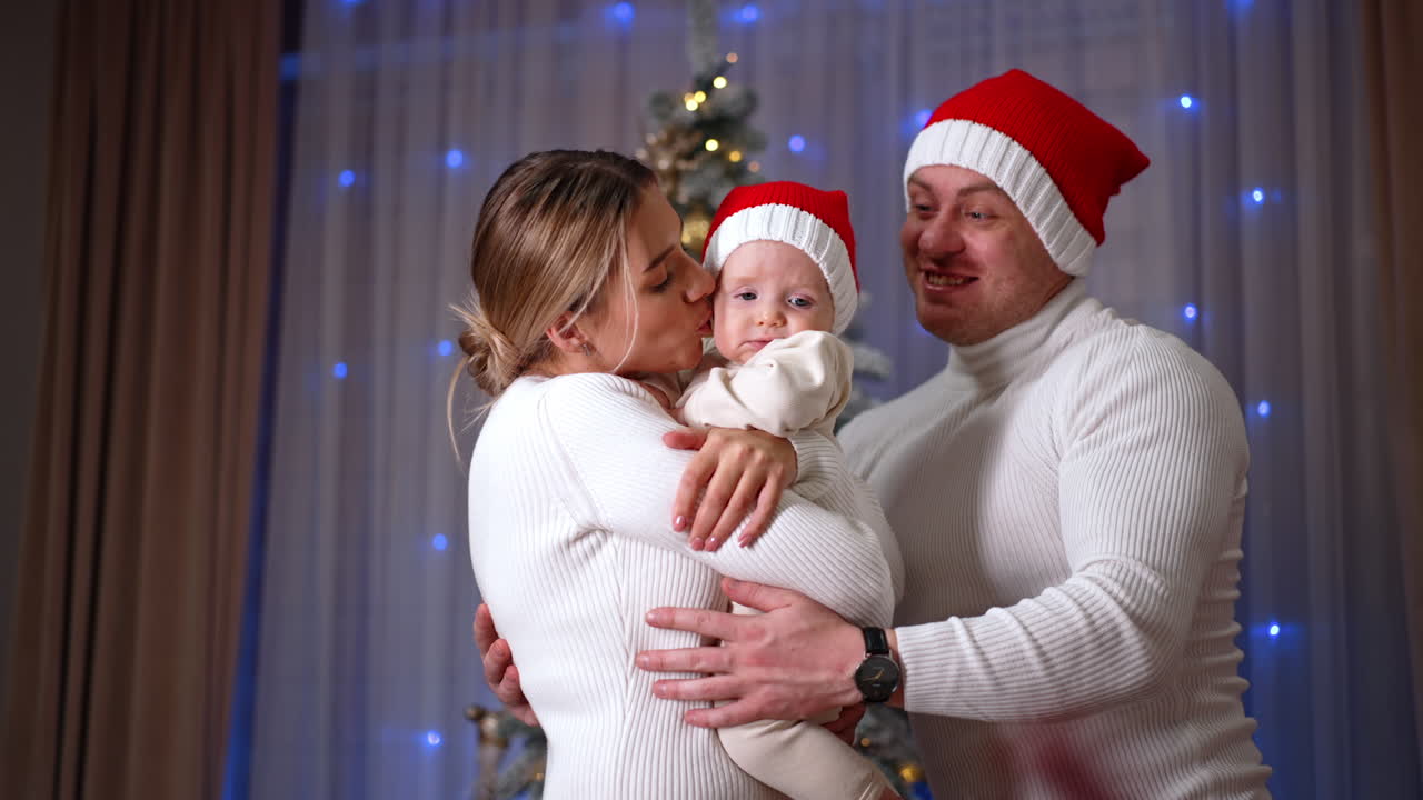 Happy mother shaking lovingly her adorable baby wearing Santa cap. Father in red cap appears and parents kiss their child. Christmas concept.