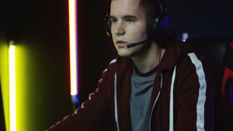 close up view of the young serious man with headphones and playing a game on the computer in a room with colorful neon lamps on the wall