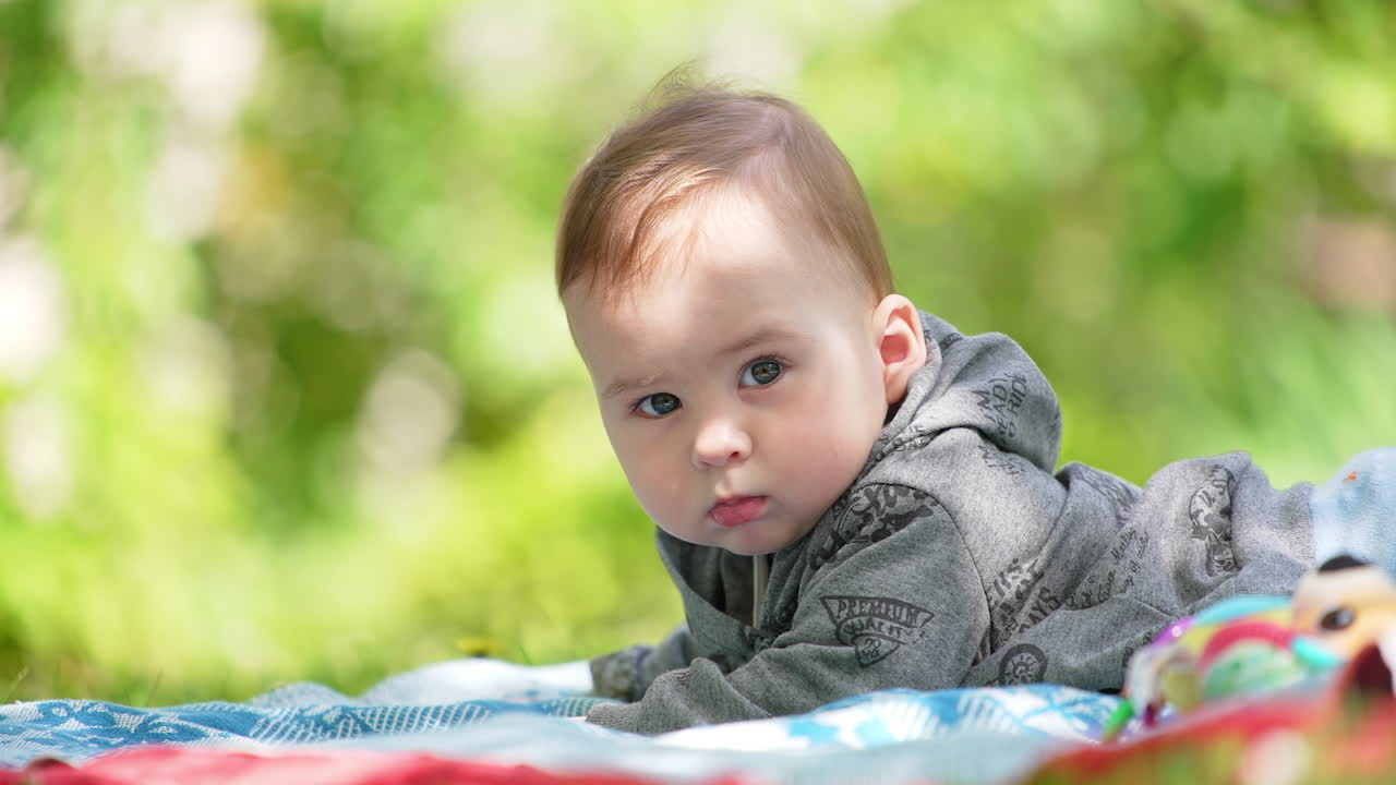 Lovely baby boy wearing grey hoody lies on the belly. Sweet kid having rest outdoors. Blurred backdrop.