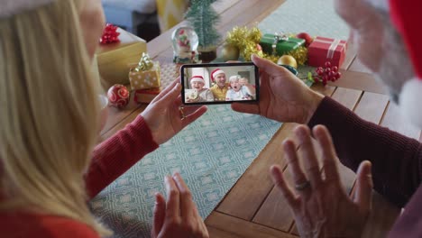 Pareja-Caucásica-Con-Sombreros-De-Santa-Usando-Un-Teléfono-Inteligente-Para-Una-Videollamada-Navideña-Con-La-Familia-En-La-Pantalla