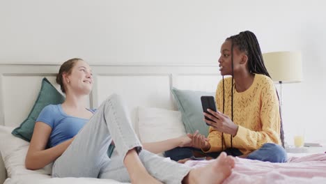 happy diverse teenage female friends lying on bed using smartphones, talking and laughing