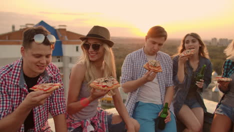 los jóvenes al atardecer en el techo disfrutan de pizza y cerveza. se sientan y hablan entre sí.