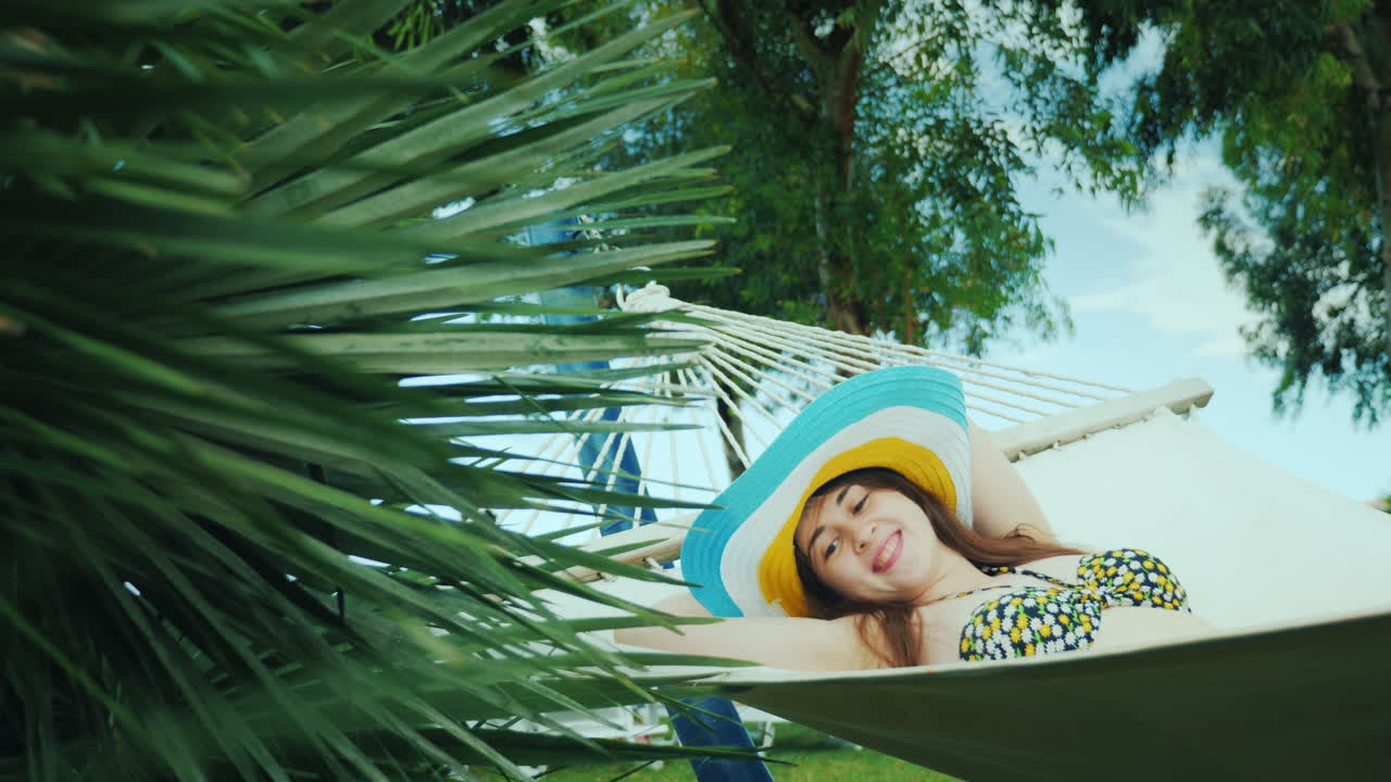 una joven feliz con un sombrero brillante de ala ancha y en bikini se balancea en una hamaca cerca de un bas abierto