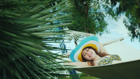 a happy young girl in a bright wide-brimmed hat and in a bikini swings in a hammock near an open bas