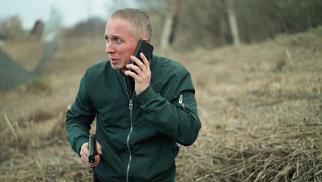 a close view of a man in a green jacket holding a handgun and speaking on a phone in a dry, grassy area, he appears tense and focused