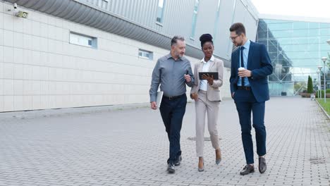 Afroamerikanische-Junge-Geschäftsfrau-Und-Zwei-Kaukasische-Geschäftsmänner,-Die-Die-Straße-Hinuntergehen-Und-Mit-Einem-Tablet-über-Die-Arbeit-Diskutieren