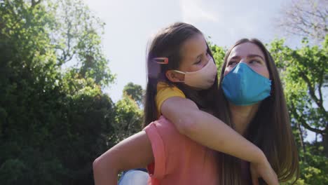 madre caucásica con máscara facial cargando a su hija en la espalda en el jardín