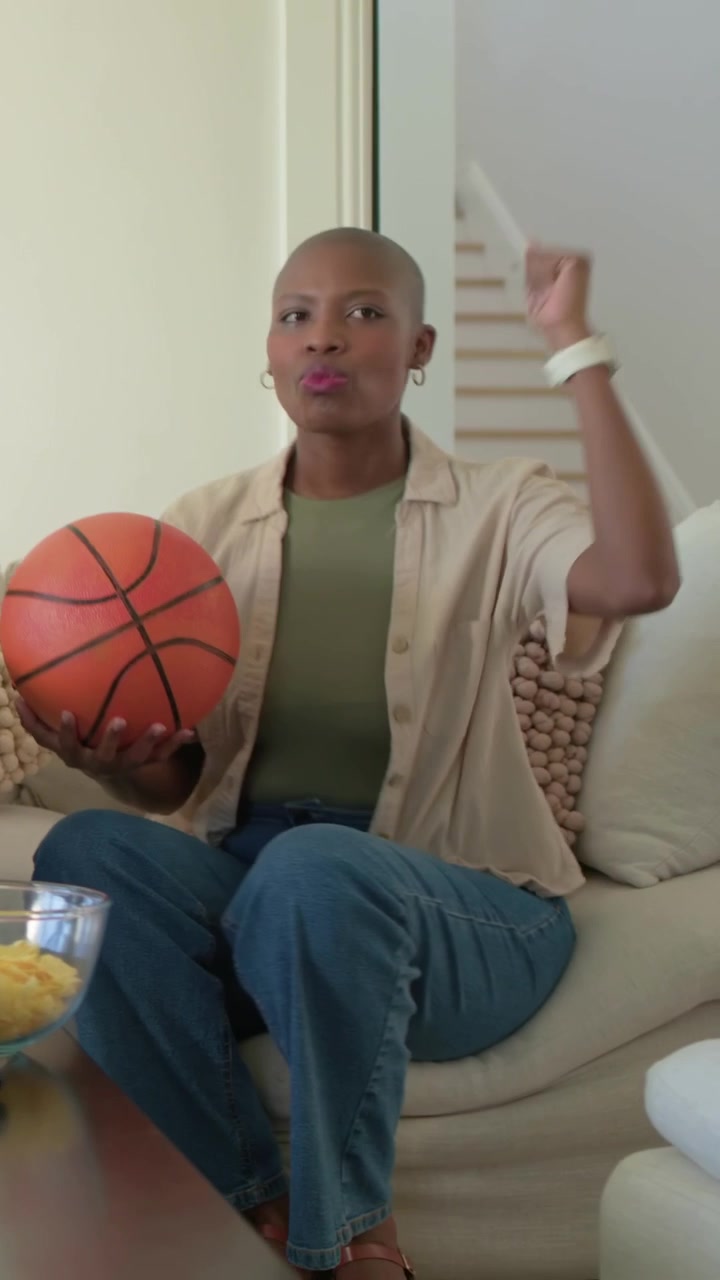 Video verticale: Sentendo un rumore, una donna con top beige e jeans tiene un pallone da basket a casa, prendendo una patatina