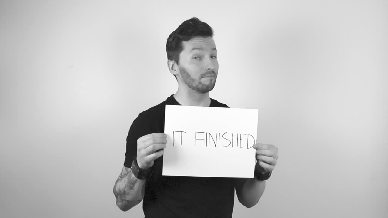 Grayscale footage of a tattooed man with short hair and trimmed beard lifts up a big white paper with the writing IT FINISHED on it with capital letters, the man is happy and is smiling about this.