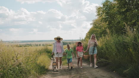 family strolling along dirt road surrounded by lush greenery under bright sky, all wearing dark sunglasses, with woman on right walking dog on leash while another carries picnic basket