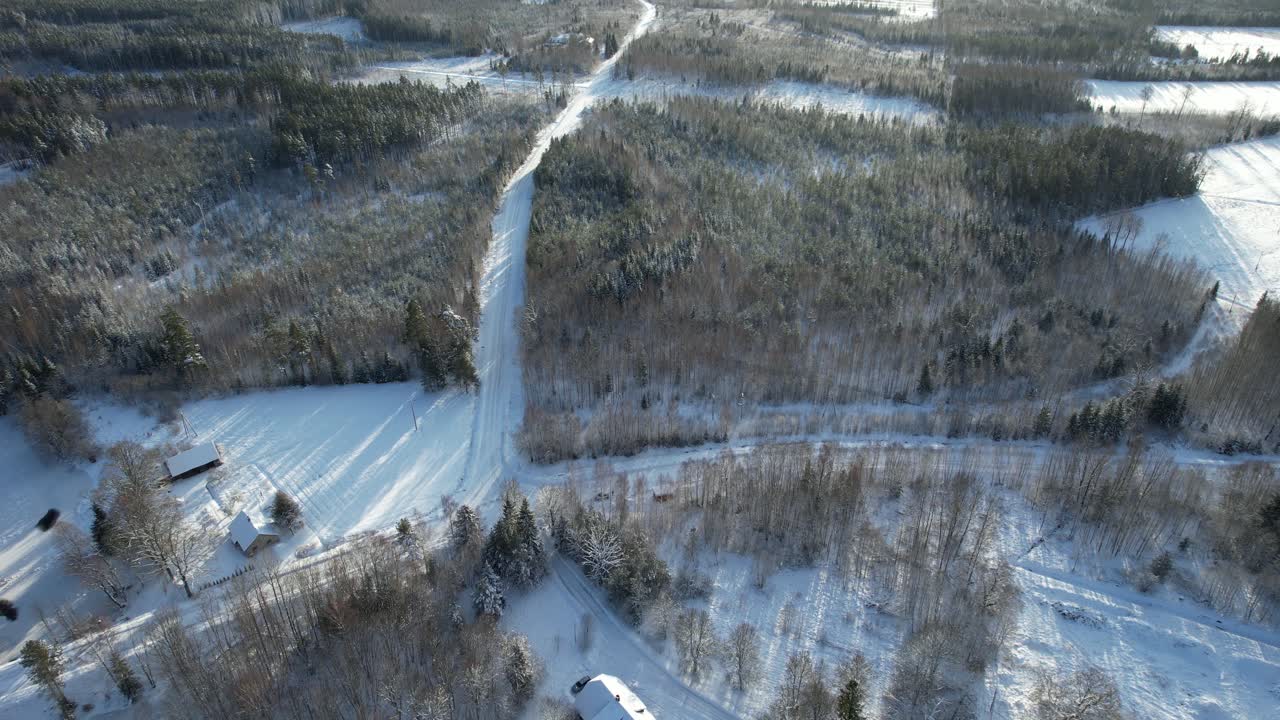 Aerial view of a serene winter countryside featuring snow-covered trees, wooden houses, and a winding road through a frosty forest.