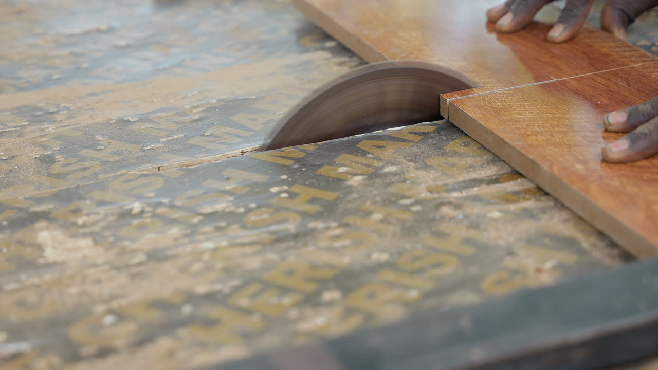 Sega da banco che taglia una tavola di legno guidata da mani di falegname scure, lama rotante che morde l'impiallacciatura, pioggia di polvere, operatore che stabilizza la tavola per l'installazione del pavimento, banco da lavoro industriale, ronzio del motore, concentrato.