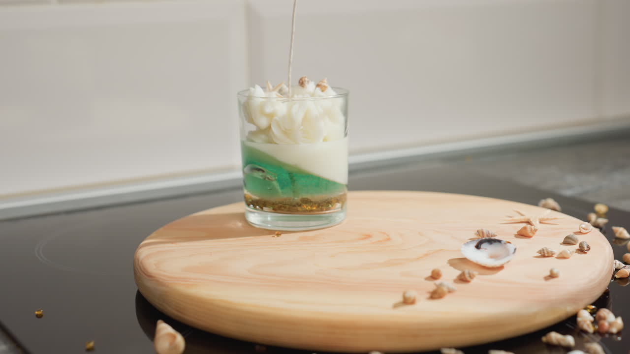 Female artist places dessert jar on decorated wooden tray with seashell embellishments scattered across tabletop in cozy kitchen setting, combining creativity and elegance with handcrafted detail