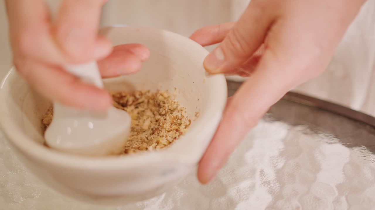 Mani che premono la base di biscotti graham per la torta di zucca, pestello in ceramica che compatta le briciole su una tovaglia di pizzo, movimento costante dello chef, vista ravvicinata di una consistenza uniforme e un piano di lavoro caldo, preparazione precisa di un dolce festivo.