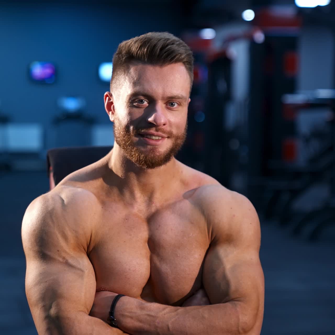 Portrait of bodybuilder indoors. Young bearded sportsman with athletic body sitting in gym. Muscular athlete without shirt. Front view.