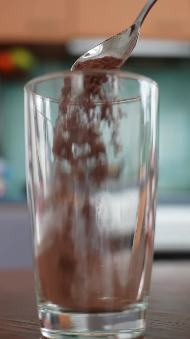 Primo piano verticale di cacao in polvere che cade da un cucchiaio in un bicchiere vuoto su un tavolo di legno, semplice passaggio in cucina per preparare una miscela per bevanda al cioccolato