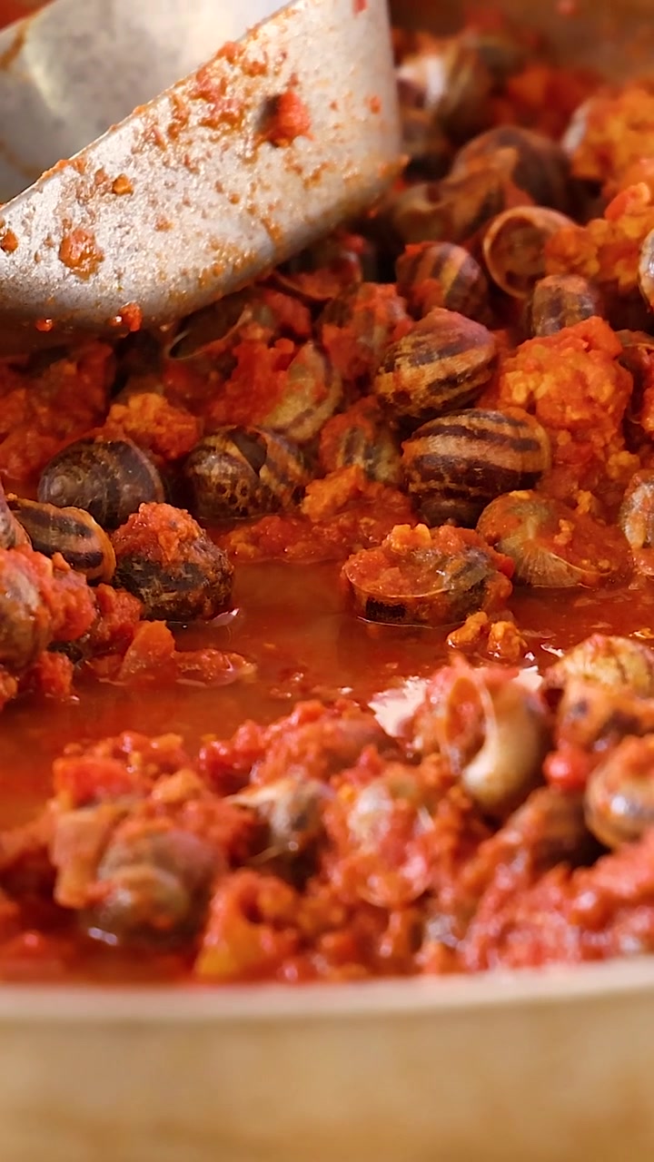 Un mestolo di metallo mescola saporite lumache escargot che sobbollono in una ricca salsa di pomodoro rossa e bollente.