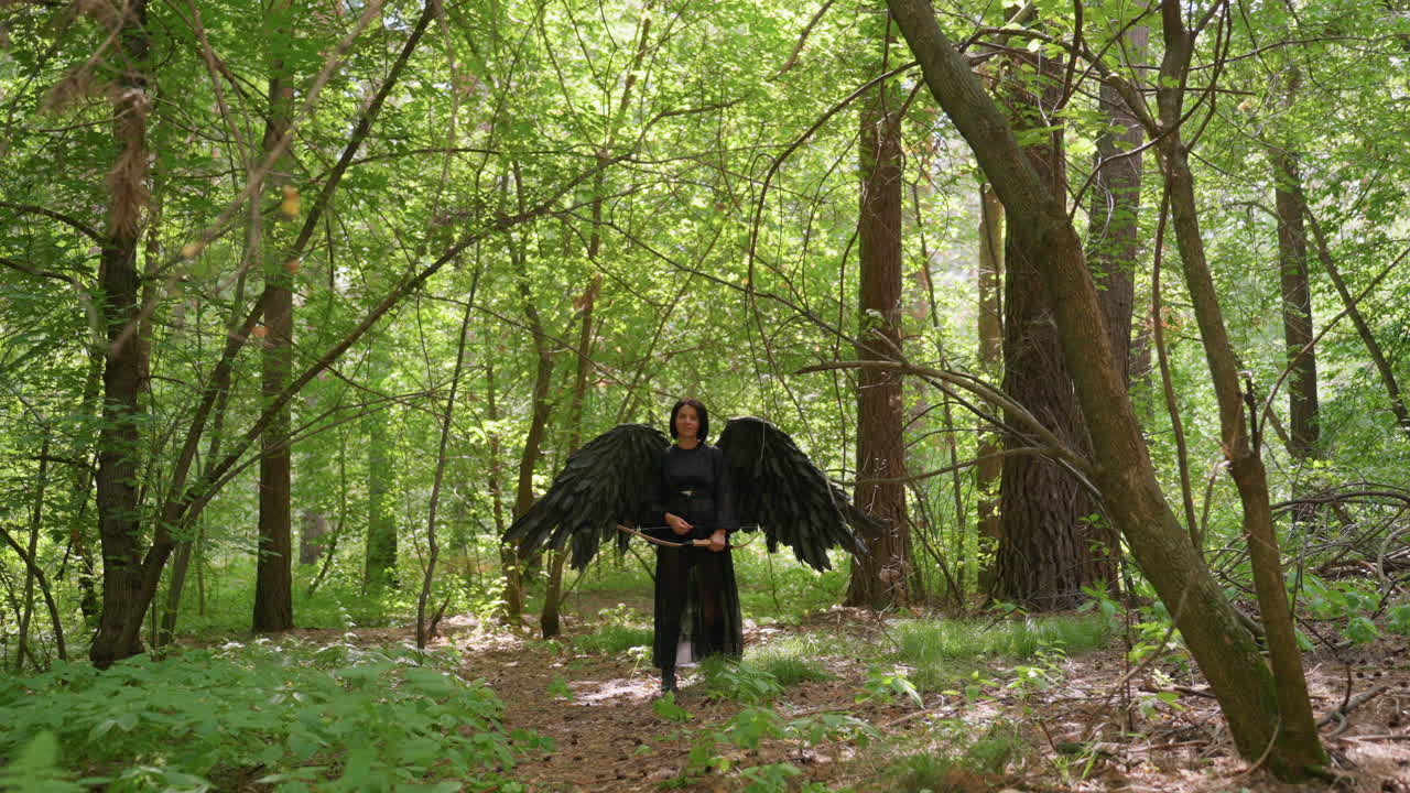 Archer goddess with black wings stands tall among forest trees lowers her bow, sunlight glowing through green leaves, symbolizing courage