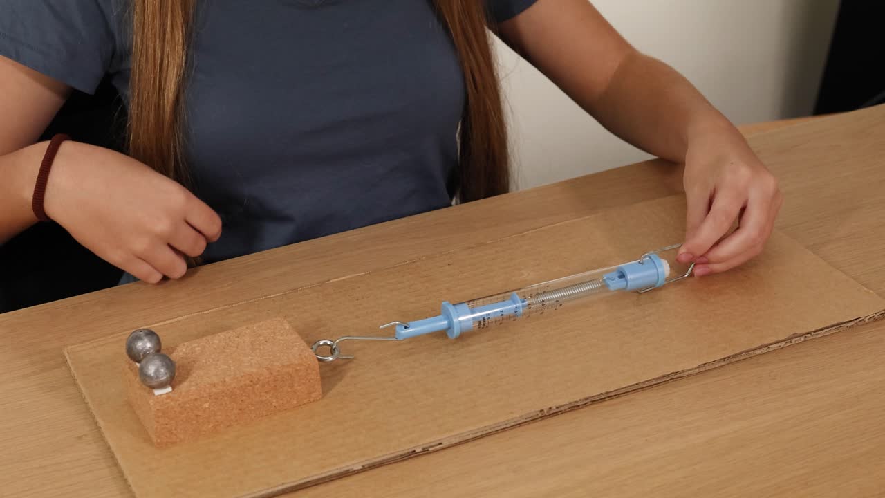 Hands manipulate a spring scale to measure force on a cardboard surface, illustrating friction and weight dynamics