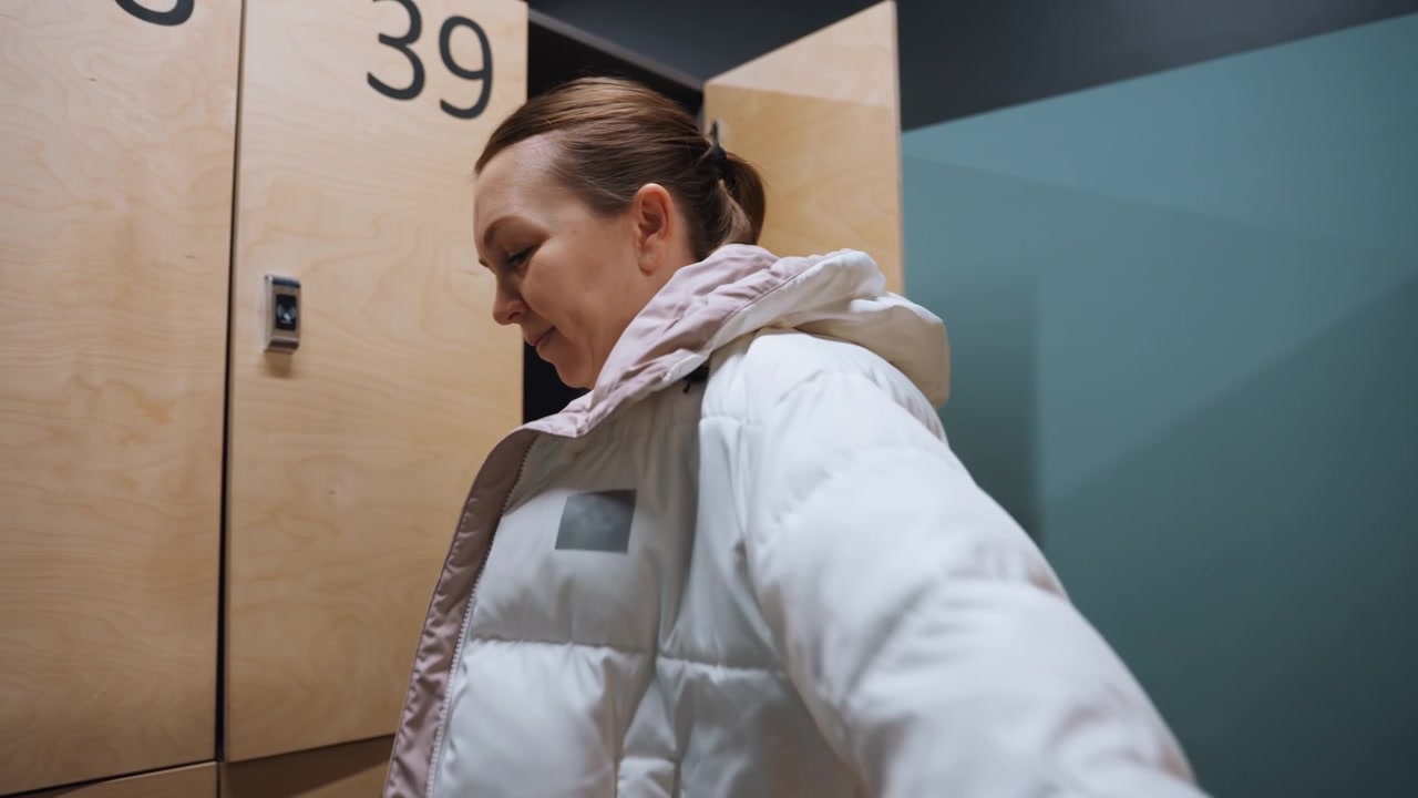 Locker room exit badminton player puts on jacket and secures bag. After practice player lifts coat from locker, zips hood, adjusts shoulders, collects key, confident relieved expression, ready