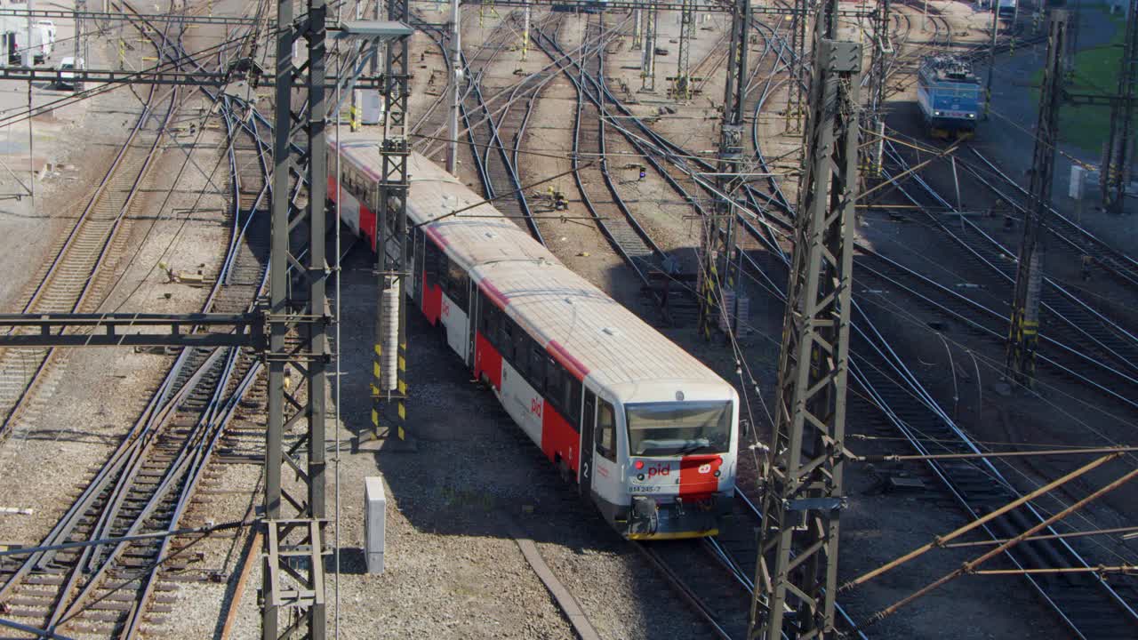 A red and white passenger train moves along railway tracks toward a major station in Prague, captured in daylight from an elevated, wide-angle perspective