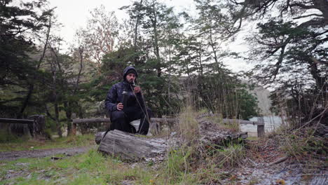 person sitting on fallen tree trunk in forest campsite area and drinking mate.