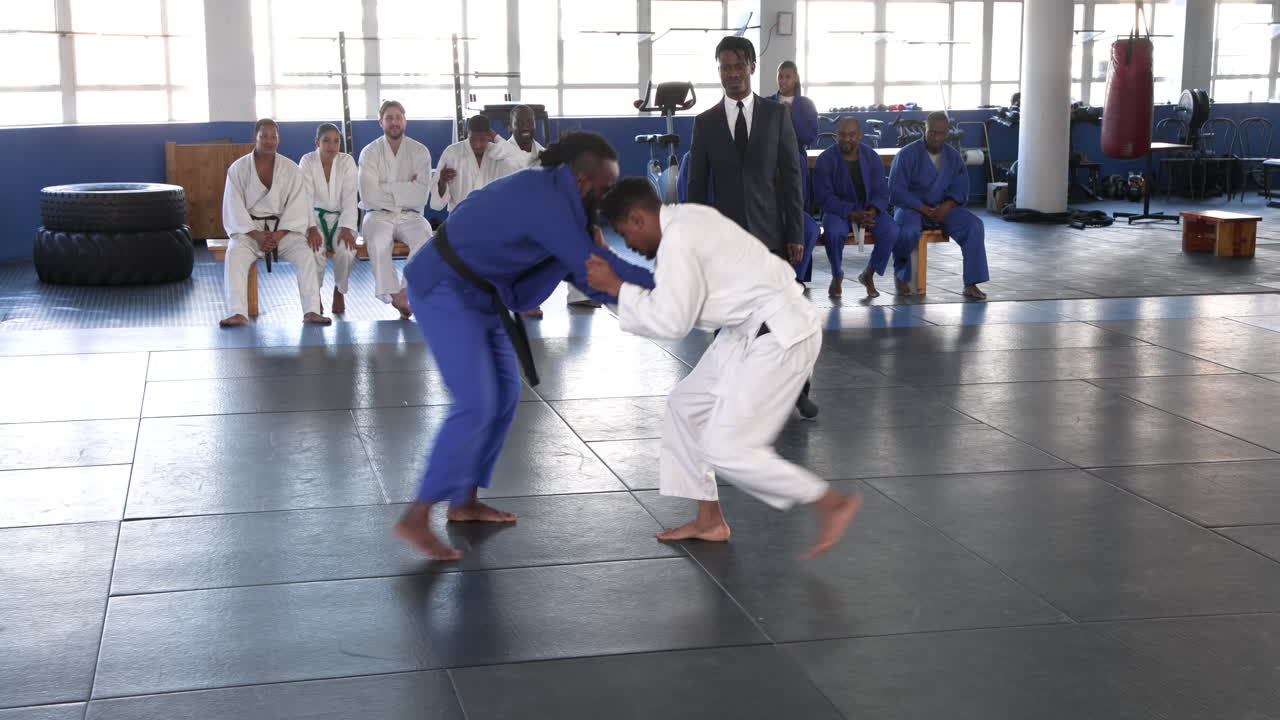 Practicing judo, athletes grappling on mat with coach and teammates watching