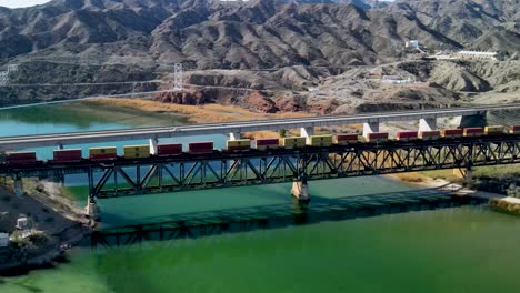 Cargo-Train,-Crossing-Steel-Bridge-Over-Colorado-River,-The-Border-Between-California-and-Arizona,-I-40-freeway-East,-Wide-Angle-Rotating-Fly-Over-Drone-Shot