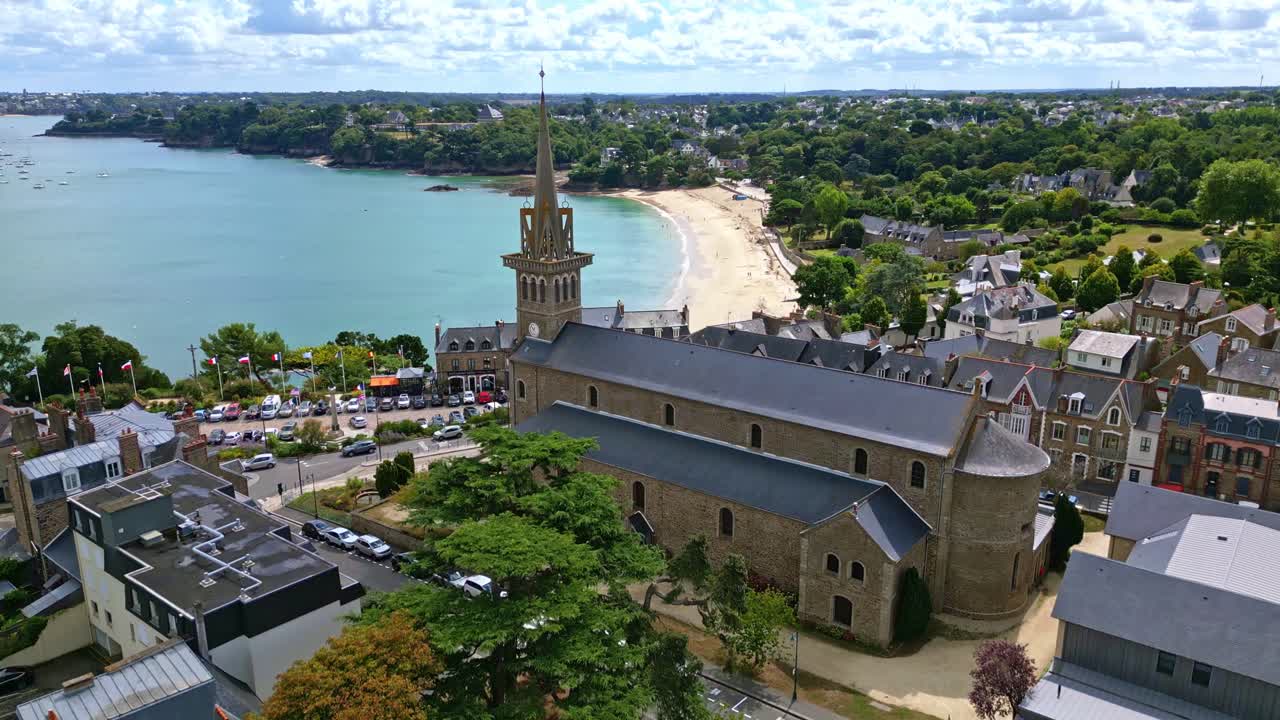 Drone ascends tilting down over Eglise Notre Dame d’Emeraude with trees, stone stairs, and coastal backdrop. Dinard, France