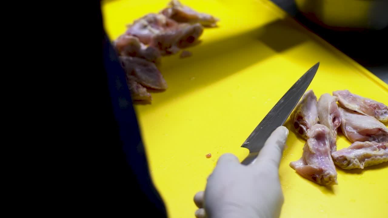 chef con guantes blancos esterilizados cortando alas de pollo con un cuchillo afilado en una tabla de corte amarilla para una receta de milanesas