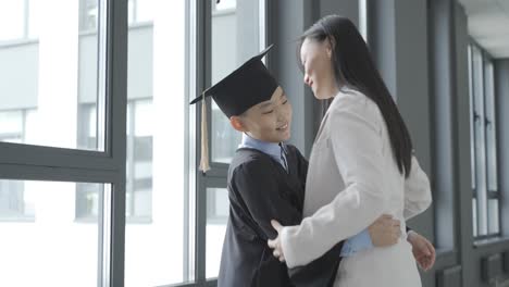 asian mom hugging her son. he's wearing a gown and a mortarboard.