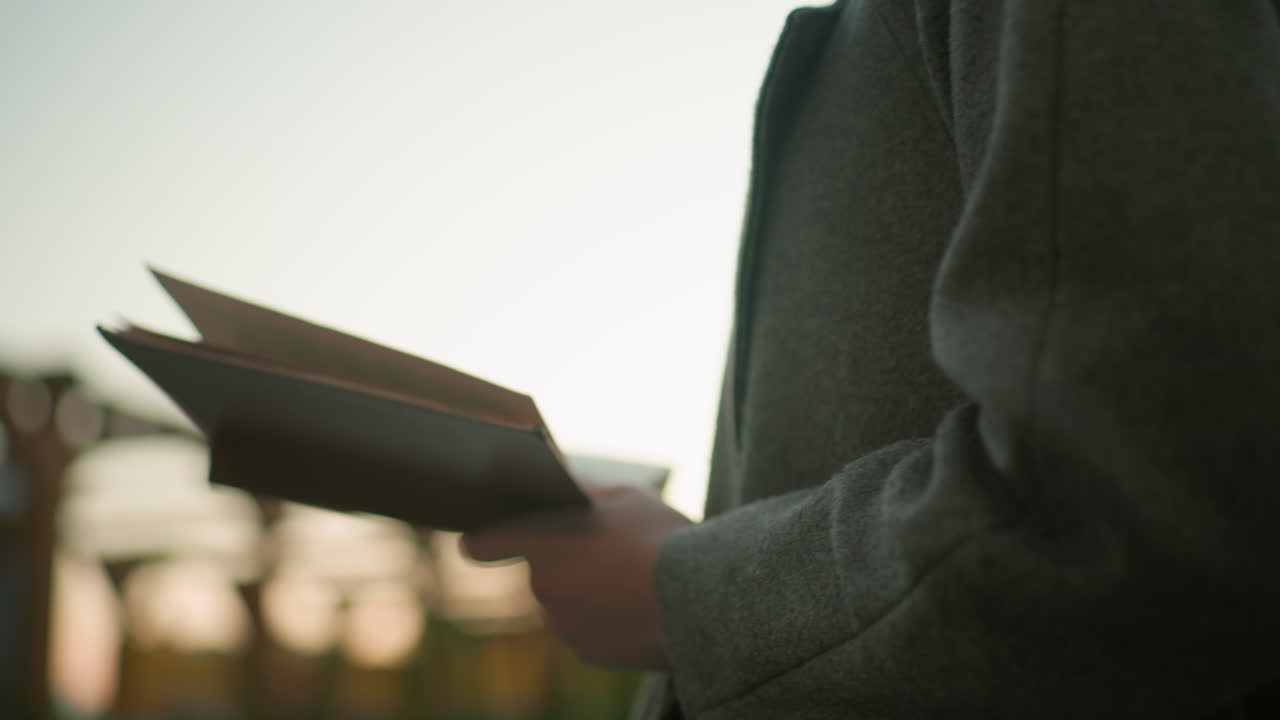 Lady with long flowing hair holds open book while standing outdoors at sunset, back view highlighting hair texture and coat detail with blurred wooden structure and city buildings