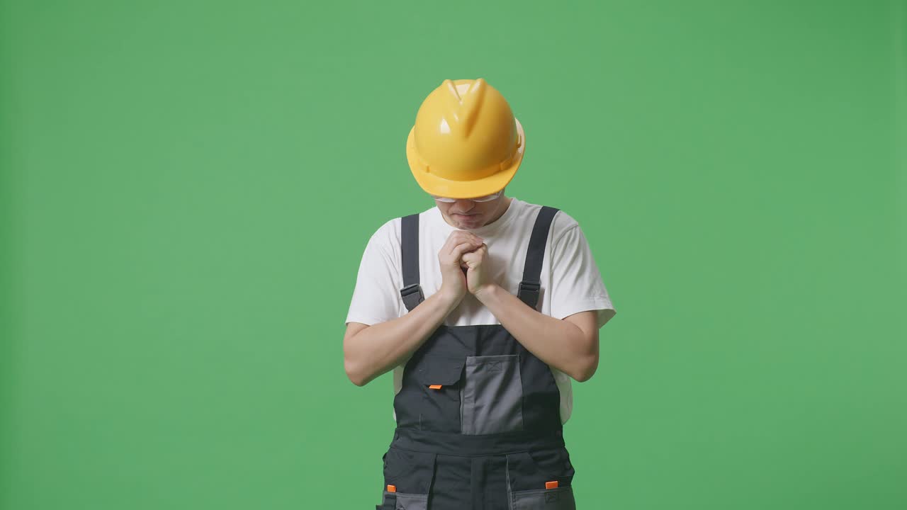 азиатский мужчина рабочий в защитных очках и защитном шлеме молится о чем-то, стоя в зеленом экране фоновой студии