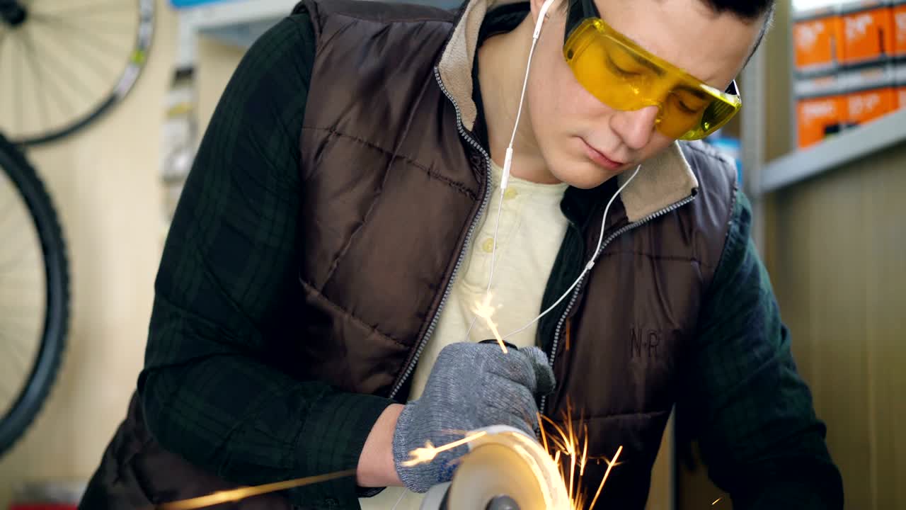 серьезный ремонтник в защитных очках использует электрическую круговую пилу при резке металлических запасных частей велосипеда на рабочем месте. яркие искры на переднем плане.