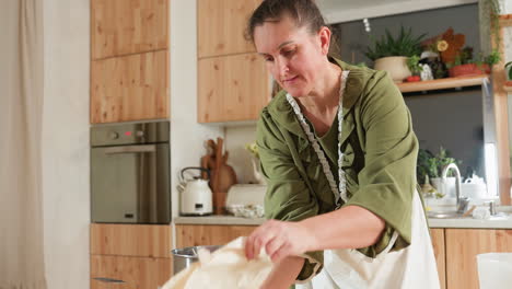 woman in green gown places white jug on countertop, swiftly picks cone-shaped cloth, skillfully inserts metallic cone inside, preparing carefully for baking activity in bright cozy kitchen area