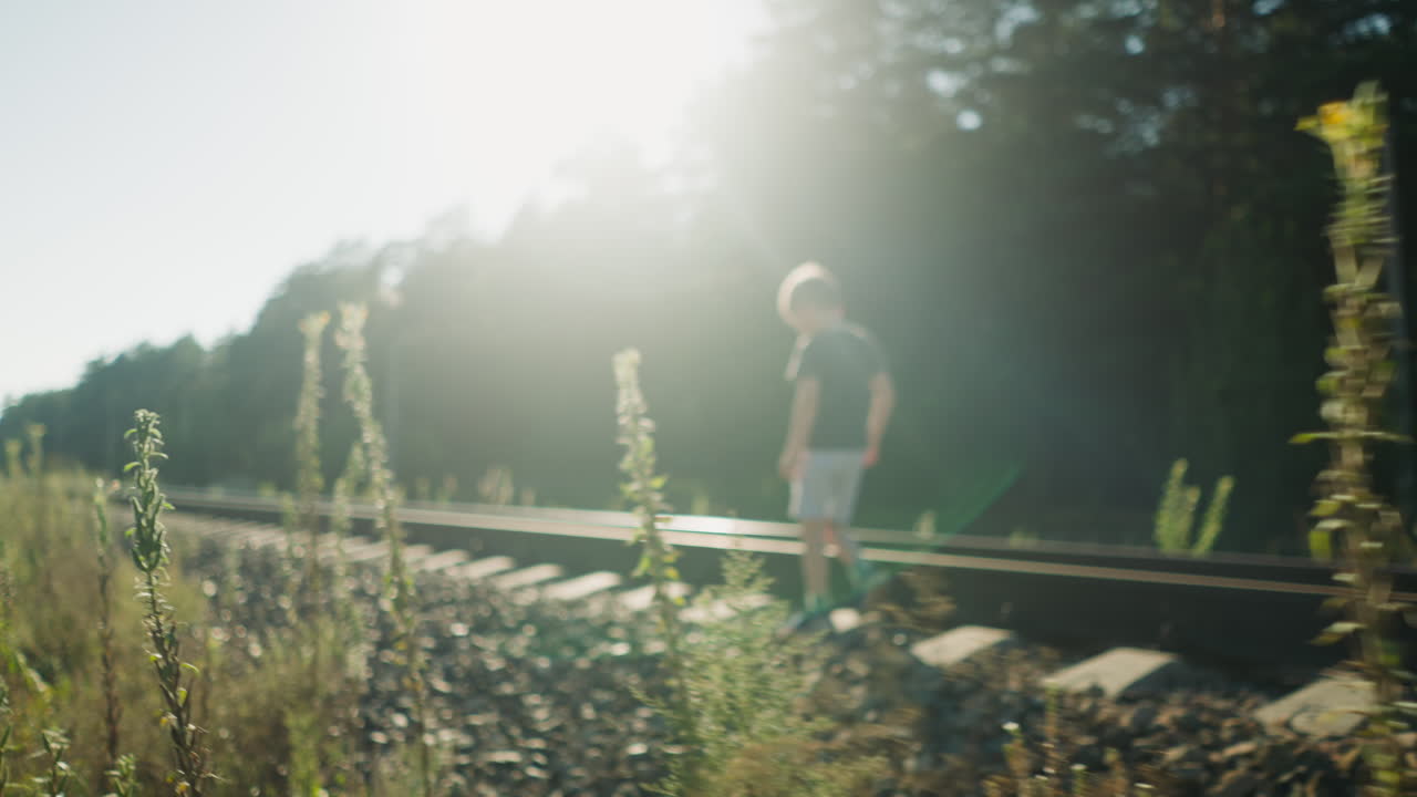 Little boy walking along rail track with bright sunlight casting strong glow through surrounding trees and wild plants, creating dramatic silhouette and peaceful rural atmosphere