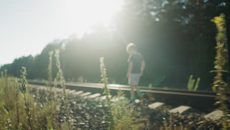 little boy walking along rail track with bright sunlight casting strong glow through surrounding trees and wild plants, creating dramatic silhouette and peaceful rural atmosphere