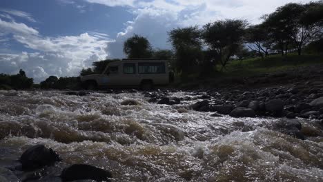 Tiro-De-Punto-De-Vista-Bajo-De-Un-Vehículo-De-Safari-Cruzando-Un-Pequeño-Rápido-De-Agua-En-Natron-Tanzania-Con-Un-Cielo-Maravilloso