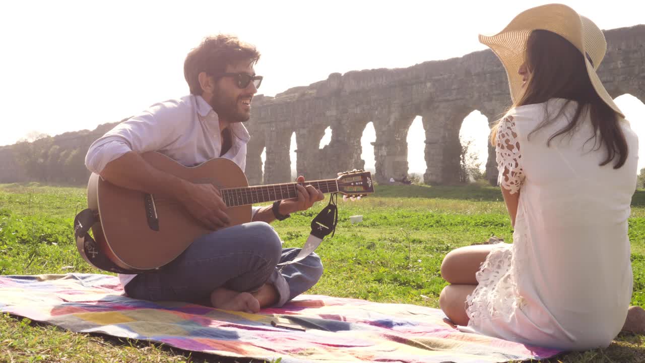 Happy young lovely couple sitting lying on a blanket in the grass in front of ancient roman aqueduct ruins in parco degli acquedotti park in rome romantic play guitar sing beautiful girl with hat bucolic slow motion