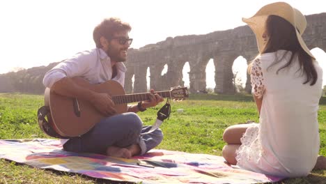 happy young lovely couple sitting lying on a blanket in the grass in front of ancient roman aqueduct ruins in parco degli acquedotti park in rome romantic play guitar sing beautiful girl with hat bucolic slow motion