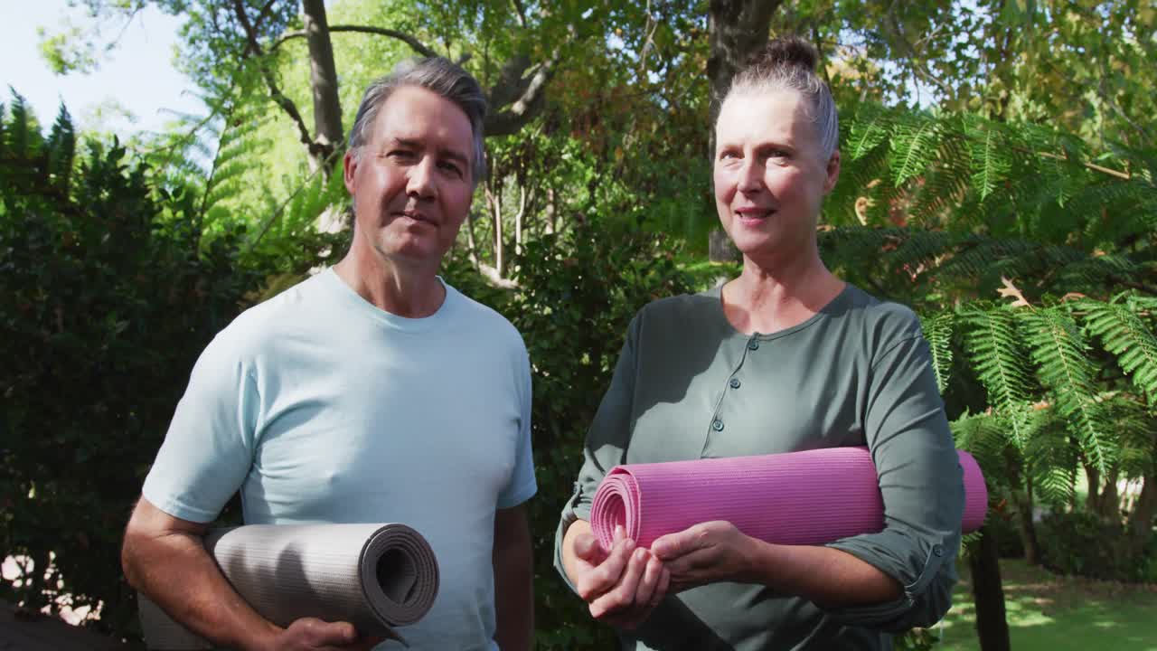 Glückliches älteres kaukasisches Paar spricht und lächelt, während es Yogamatten im Garten hält.