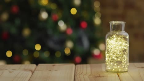 video de una mesa de madera con luces de hadas en un frasco sobre un árbol de navidad desenfocado con bolas