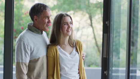 Sonriente-Padre-Caucásico-Y-Su-Hija-Adolescente-Abrazándose-Y-Mirando-Por-La-Ventana