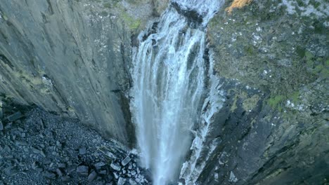 Top-down-view-of-waterfall-cascading-over-icy-steep-cliff-to-frosted-dark-stony-beach-below