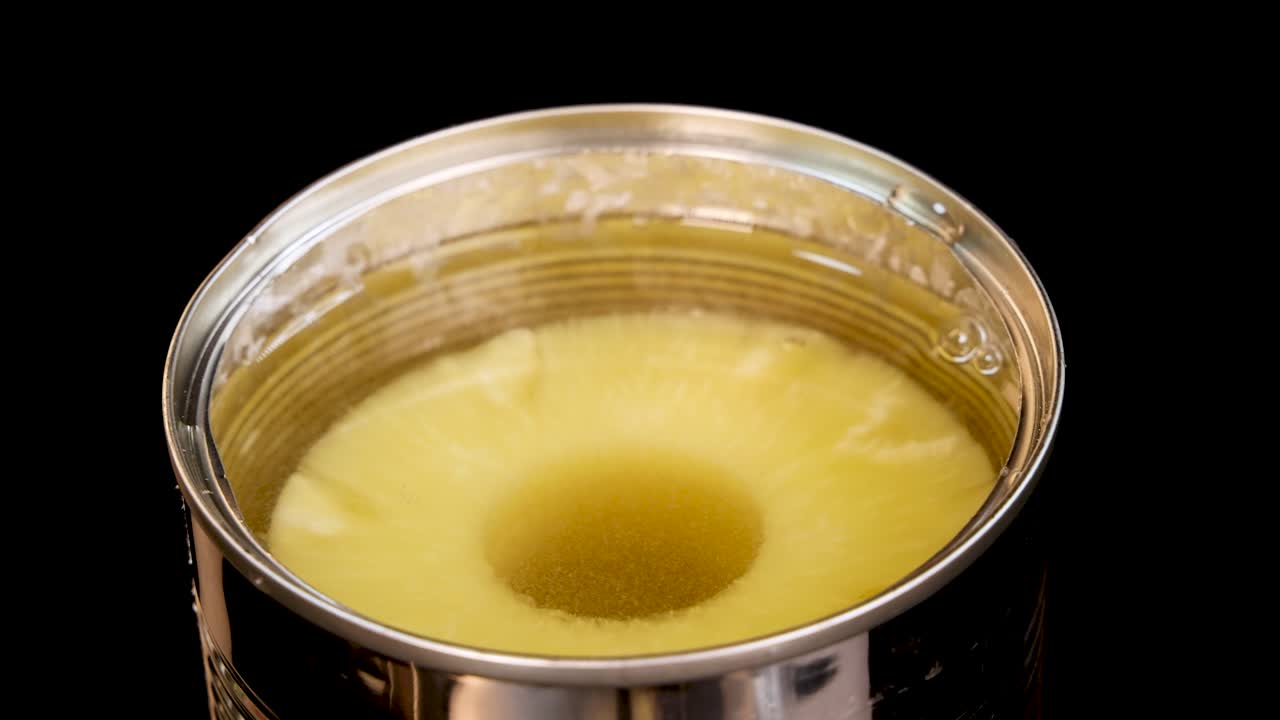 A pineapple ring is dropped into a metal can of syrup, creating a splash and ripples. Studio lighting, overhead view, black background