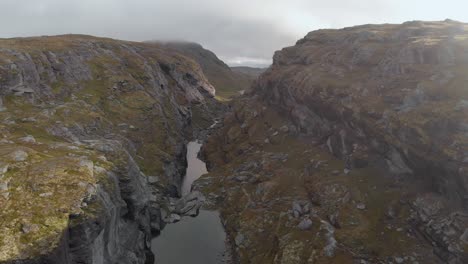 River-flowing-below-steep-canyon-with-dark-clouds,-Norway-highlands,-aerial-view