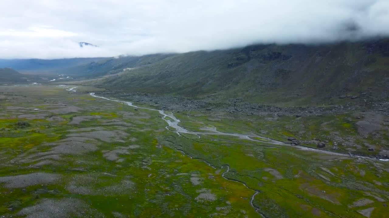 Aerial drone footage view flying over Sweden rocky landscapes with narrow river flowing on the green moss covered rocks during a cloudy day. Large mountain visible in the background and white clouds.