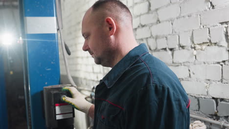 mechanic in blue uniform wearing protective gloves presses lift button in garage, background features brick wall, blue lift, and bright workshop lighting creating professional repair environment